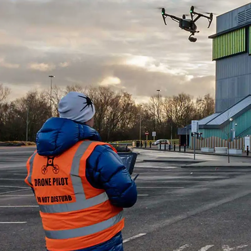 A man in an orange vest is observing an aerial drone flying over a building.